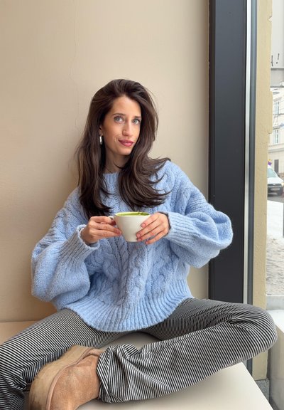 Mujer con cabello largo y castaño, vestida con un suéter de punto azul y pantalones a rayas, sentada con las piernas cruzadas junto a una ventana, sosteniendo una taza de té verde o matcha.