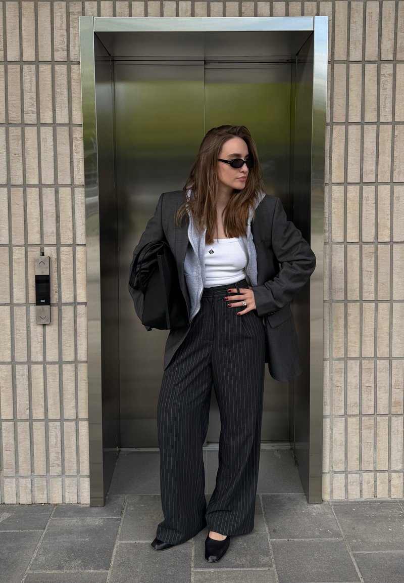 Femme portant des lunettes de soleil et un blazer superposé avec un sweat à capuche, debout dans une porte d'ascenseur fermée, tenant un sac noir contre un mur carrelé.