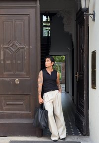 Uomo con tatuaggi che indossa una maglietta nera senza maniche e pantaloni bianchi si trova in una porta tenendo una borsa nera, guardando a sinistra.