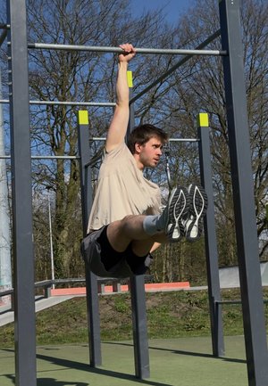 Una persona realiza un ejercicio de suspensión en una estructura metálica gris. Lleva una camiseta ligera y pantalones cortos, con zapatos deportivos. Se pueden ver árboles de fondo.