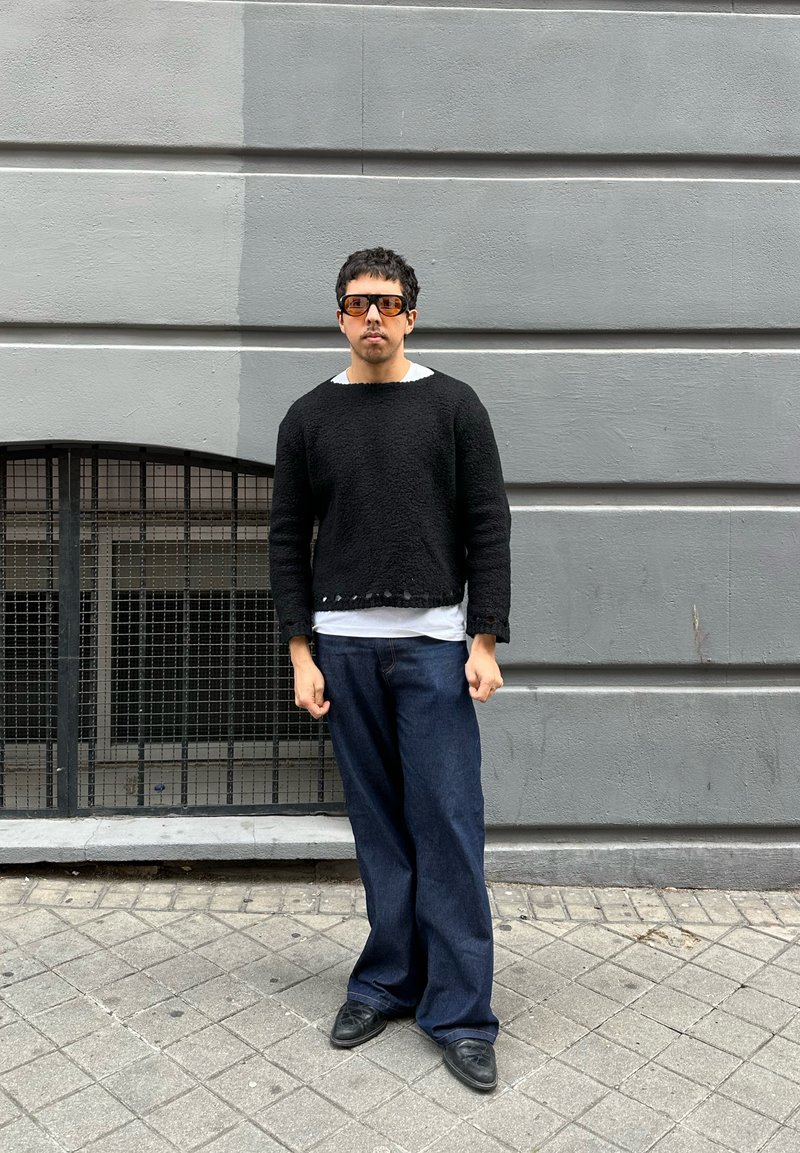 Mann trägt schwarzen Pullover, weißes Hemd, weite blaue Jeans und braune Sonnenbrille, steht auf einem gefliesten Gehweg neben einer grauen Mauer mit vergittertem Fenster.