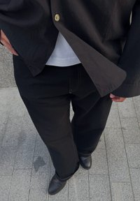 Schwarzes Hemd über einem weißen T-Shirt, kombiniert mit schwarzen weiten Hosen und schwarzen Knöchelstiefeln, auf einem strukturierten grauen Pflaster.