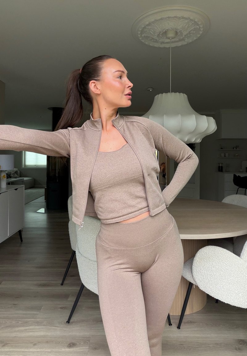 Woman in beige matching activewear stretching in a modern dining room with a round wooden table and white chairs.