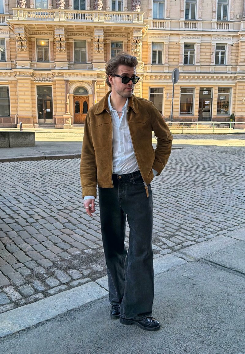 Homem vestido com casaco castanho, camisa branca, calças pretas de perna larga e óculos de sol, a pé numa rua de calçada com um edifício histórico ao fundo.