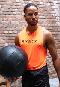 Fekete medicinlabda, amelyet egy narancssárga ujjatlan sporttopot viselő személy tart a vállának magasságában, rajta fekete "HYROX" felirattal, egy téglafal előtt.