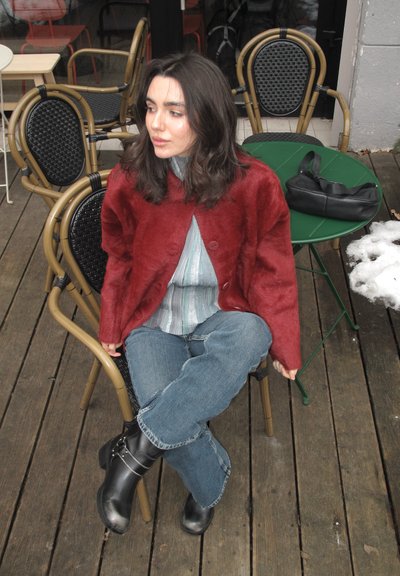 Mujer de cabello oscuro con chaqueta roja y jeans azules sentada con las piernas cruzadas en una silla de mimbre en una mesa de café de madera, con una bolsa negra cerca.