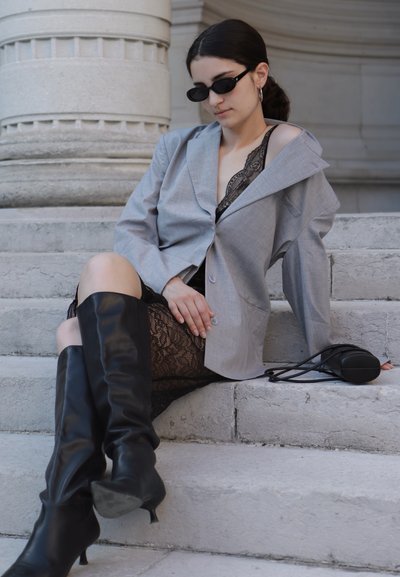 Blazer oversized de color gris claro sobre un vestido de encaje negro, combinado con botas de cuero negro hasta la rodilla. Los accesorios incluyen pequeñas gafas de sol negras.