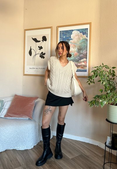 Suéter de punto color crema con escote en V y mangas cortas; falda corta negra; botas negras hasta la rodilla; decoración ligera con obras de arte enmarcadas y una planta.