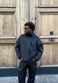 Manteau en laine grise avec col montant et détails de boutons, accompagné d'un pantalon en cuir noir. Une porte en bois en arrière-plan ajoute de la texture.