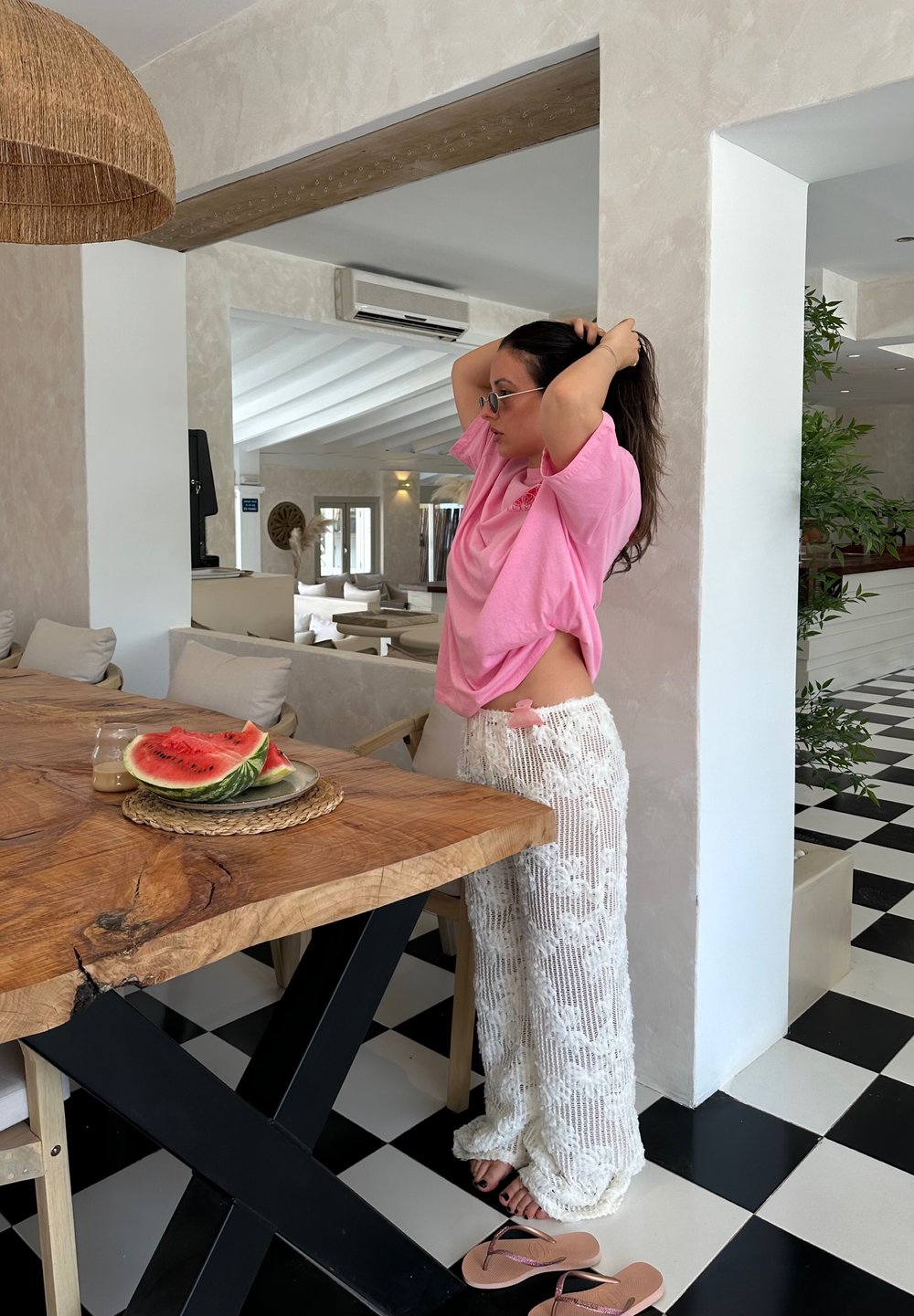 Ροζ oversized t-shirt, λευκά υφαντά παντελόνια με λουλούδια, ξύλινο τραπέζι με φέτες καρπουζιού, μαύρο και λευκό καρό πάτωμα.
