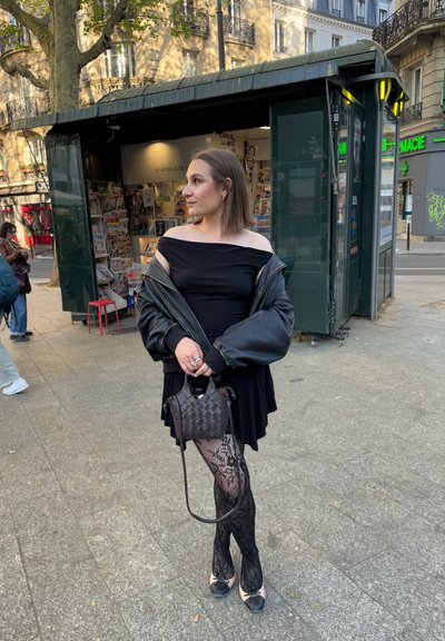 Vestido negro de hombros al descubierto, chaqueta bomber negra de gran tamaño, medias de encaje y tacones negros. Bolso de mano tejido marrón. Entorno urbano con quioscos.