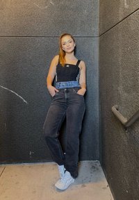 Body preto justo com riscas laterais brancas, jeans pretos de cintura alta e sapatilhas cinza claro. O cenário inclui uma parede texturizada cinza escuro.