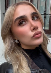 Portret osoby z długimi, jasnoblond włosami, ubraną w czarny top z kołnierzykiem i brązową kurtkę, z dużymi perłowymi kolczykami i wyraźnym makijażem.