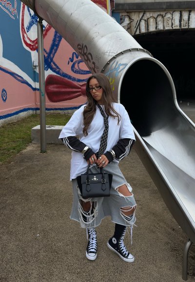 Joven mujer que lleva shorts rotos, corbata larga, zapatillas negras de caña alta y gafas, está de pie sosteniendo un bolso negro cerca de un gran tobogán de metal al aire libre.