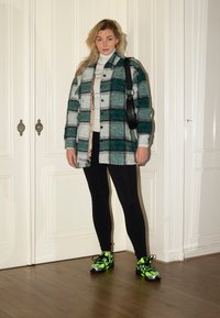 Veste chemise en flanelle à carreaux verts et gris, leggings noirs, et baskets néon vert vif avec des accents sombres. Col roulé beige en dessous.