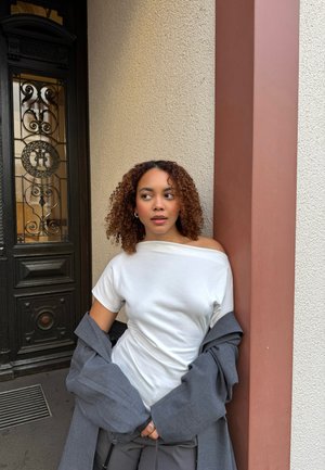 Weißes Off-Shoulder-T-Shirt kombiniert mit einem lockeren grauen Blazer und schwarzen Hosen; eine strukturierte Wand und eine dekorative Tür bieten einen kontrastierenden Hintergrund.