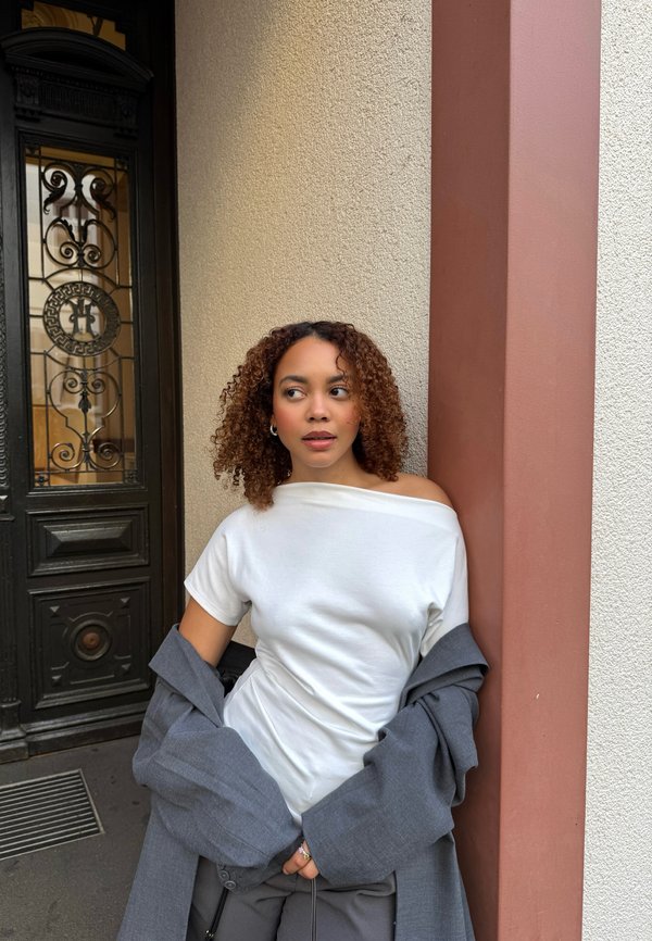Camiseta blanca con hombros al descubierto combinada con un blazer gris suelto y pantalones negros; una pared texturizada y una puerta decorativa ofrecen un fondo contrastante.