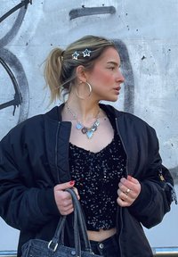 Μαύρο bomber jacket πάνω από ένα μαύρο κρουαζέ τοπ με παγιέτες, ασημένιο κολιέ σε σχήμα καρδιάς, μεγάλα σκουλαρίκια κρίκοι, κρατώντας μια γκρίζα τσάντα, με αστερόσχημες τσιμπιδάκια μαλλιών.