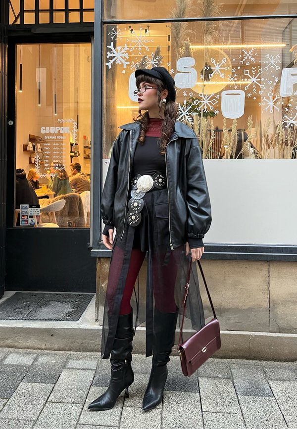 Kobieta w czarnej skórzanej kurtce, berecie, okularach i wysokich butach, stojąca przed kawiarnią ozdobioną zimowymi płatkami śniegu na oknie.