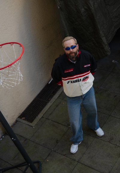 Chaqueta de la marca FUBU en blanco y negro con acentos rojos, combinada con jeans azul claro y zapatillas blancas. Canasta de baloncesto al fondo.