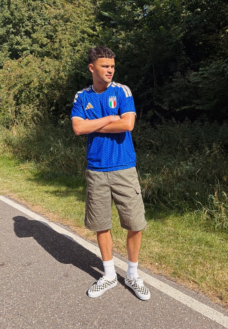 Jeune homme portant un maillot de football italien bleu, un short beige et des baskets à carreaux, debout les bras croisés au bord de la route près d'un espace vert.