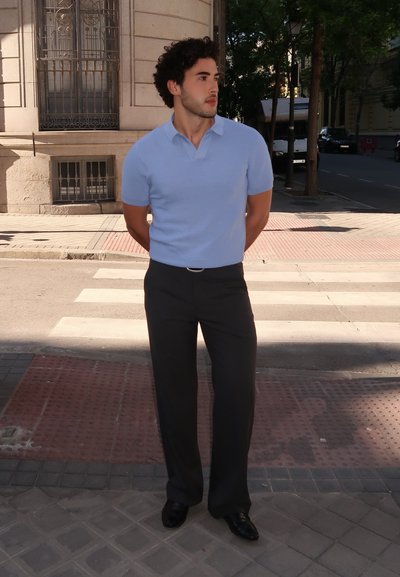 Polo de manga corta azul claro con patrón texturizado, combinado con pantalones gris oscuro y zapatos negros, de pie en una acera de la ciudad.