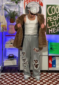 Faux-Fur brauner Mantel, weißes Tanktop, graue zerrissene Jogginghose, silberne Sneakers. Schwarze Sonnenbrille. Innenset mit Pflanzen und Kunstwerken.