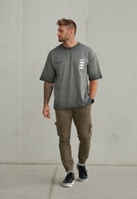 Mann med tatoveringer som bærer en grå "True Self" t-skjorte, brune cargo-bukser og svarte joggesko, som går mot en enkel lysegrå vegg.