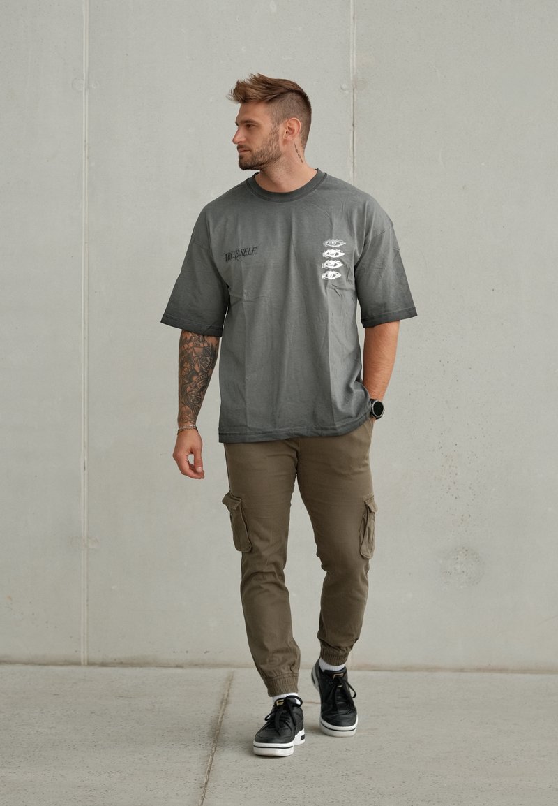 Hombre con tatuajes que lleva una camiseta gris "True Self", pantalones cargo marrones y zapatillas negras caminando frente a una pared lisa de color gris claro.