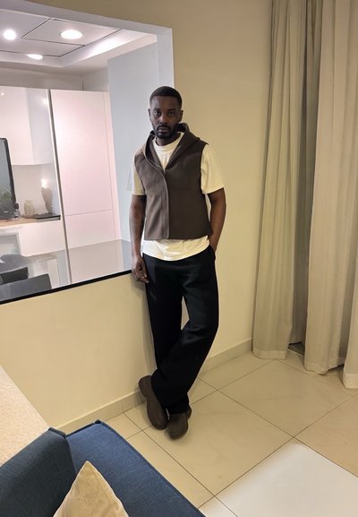 Chaleco sin mangas marrón sobre una camiseta blanca, pantalones negros holgados y zapatos marrones. Fondo neutro con luz suave y una cocina moderna.