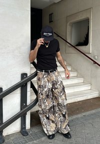 T-shirt preta de manga curta, calças largas em padrão camuflado em tons de bege e castanho, boné preto, correntes com cadeado, botas pretas.