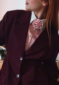 Femme portant un blazer bordeaux foncé et une chemise rose avec un broche en tissu en forme de rose, regardant sur le côté.