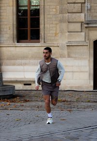 Tetovált férfi fut a szabadban, egy levelekkel borított térkövezett területen, egy nagy ablakos kőépület közelében.