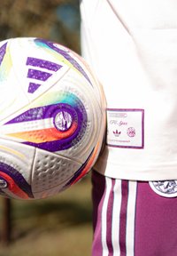 Minge de fotbal albă cu vârtejuri colorate, accente purpurii și suprafață texturată, ținută împotriva unei cămăși albe cu logo-ul AFC Ajax și pantaloni scurți purpurii cu dungi.