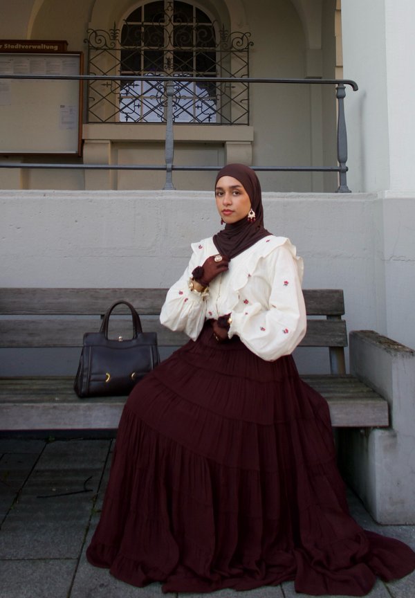 Brun tiered maxi nederdel med en cremefarvet ruffled bluse med blomsterbroderi. Tilbehør inkluderer et brunt hijab og en håndtaske.