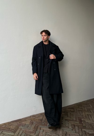 Joven con cabello rizado se apoya contra una pared beige, vistiendo un abrigo largo negro, pantalones anchos negros, zapatos negros y anillos.