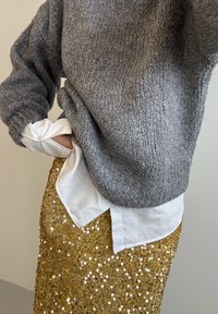 Sivi pleteni pulover z rebrastimi manšetami, nošen čez belo srajco s spodnjimi gumbi, v kombinaciji z bleščečo zlato bleščicasto krilo. Prikazuje večplastne teksture.