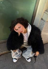 Svart, bustete pelsjakke over en grå hettegenser, kombinert med slitte, vide lyse denimjeans og svarte joggesko. Sittende mot en grønn vegg.