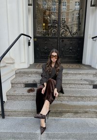 Model zittend op stenen treden, dragend een zwarte oversized blazer, bruine fluwelen broek en donkere hoge hakken met een strikdetail.