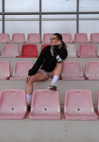 Tenue de sport composée d'un t-shirt à manches longues noir, d'un short beige, de chaussettes blanches et de baskets, assis sur des sièges en plastique rose dans un stade.