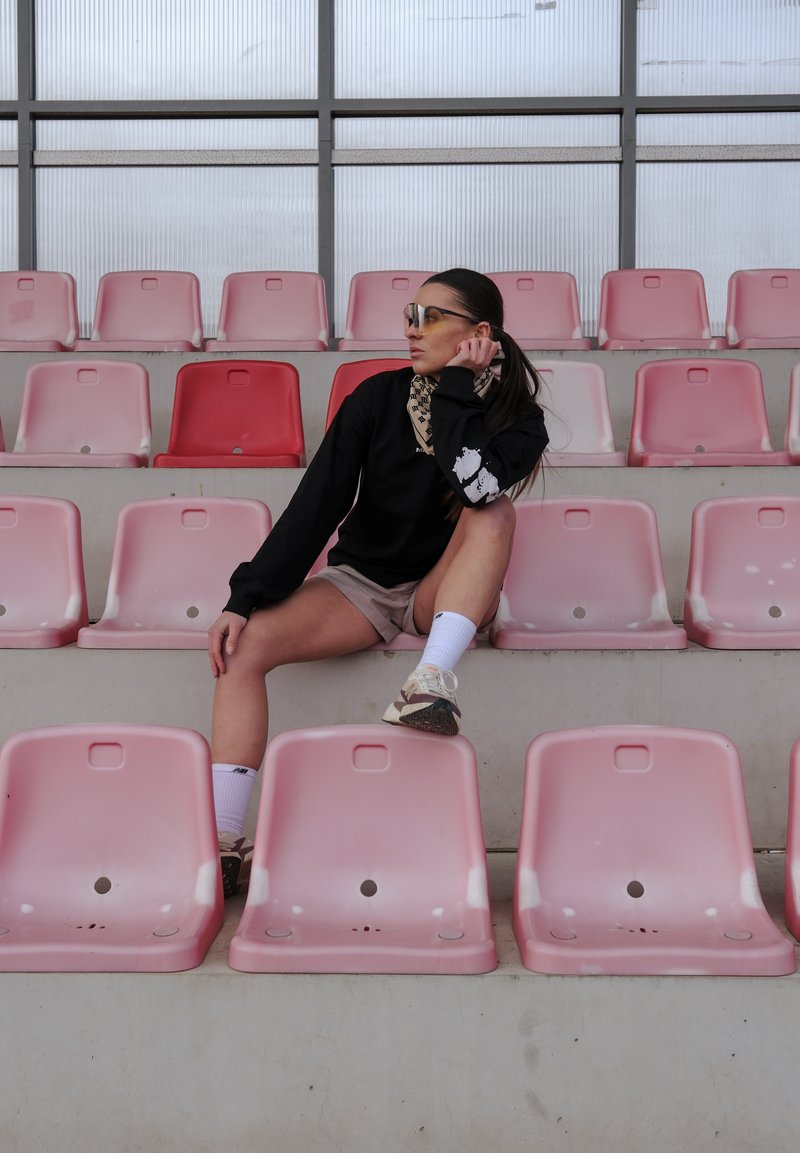 Tenue de sport composée d'un t-shirt à manches longues noir, d'un short beige, de chaussettes blanches et de baskets, assis sur des sièges en plastique rose dans un stade.