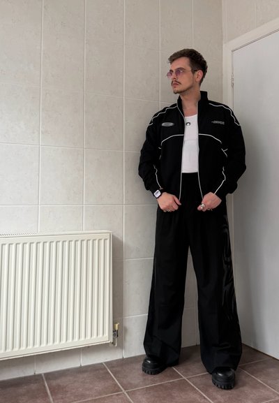 Hombre con pantalones negros de pierna ancha y chaqueta negra con ribete blanco, de pie dentro de una habitación cerca de un radiador blanco y una puerta, lleva gafas de sol y anillos.