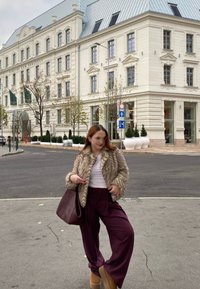 Ψεύτικη γούνα μπουφάν, λευκό τοπ και μπορντό ριγέ παντελόνι σε φαρδιά γραμμή. Καφέ μπότες μέχρι τον αστράγαλο και μία μεγάλη σκούρα τσάντα. Φόντο από αστική οδό.