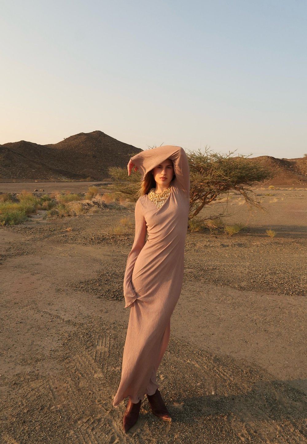 Kvinna i lång beige klänning och stövlar poserar med armen upplyft i ett ödsligt ökenlandskap med kullar och glest växtliv.