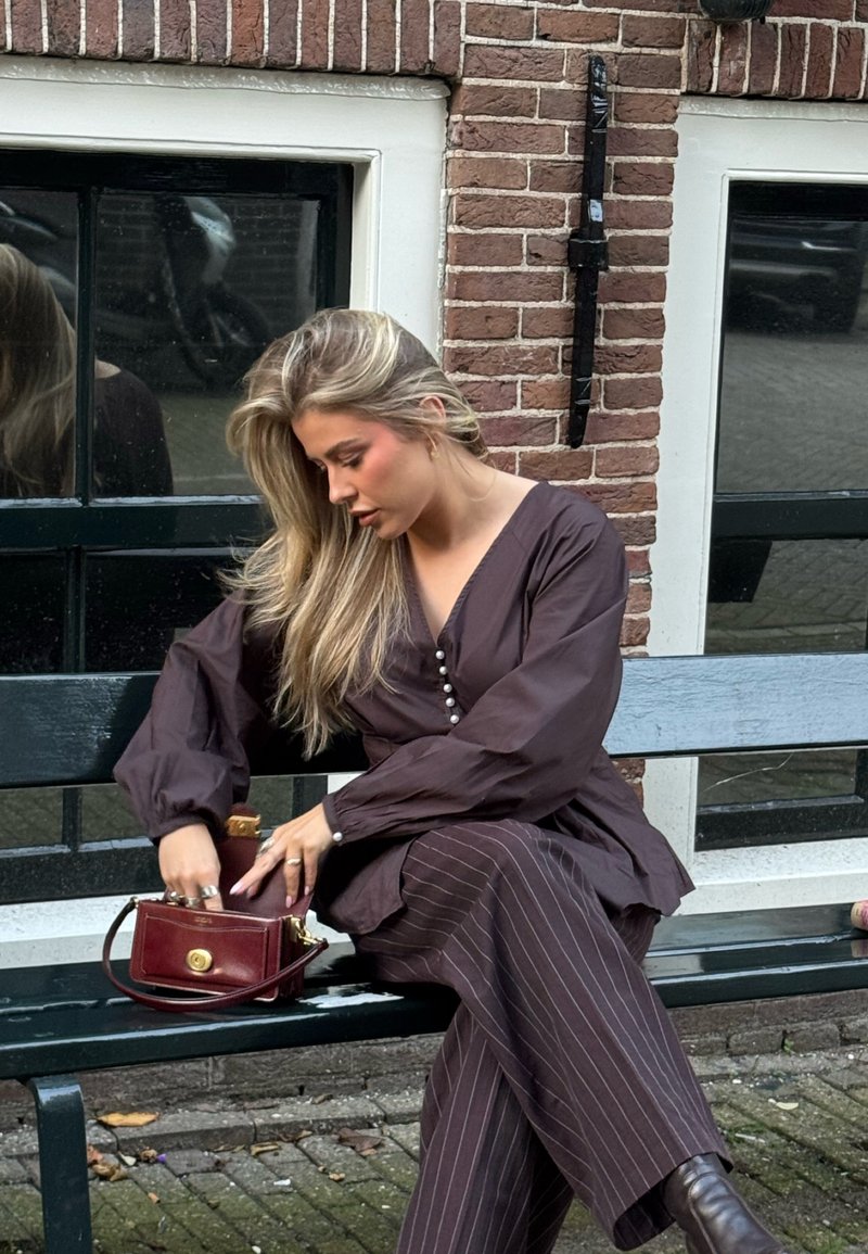 Vrouw met lang blond haar, gekleed in een donkere blouse en gestreepte broek, zittend op een bankje buiten, reikend in een kleine bruine handtas.
