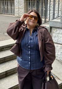 Hnědá, nadměrná bunda přes námořnickou modrou, pruhovanou košili s knoflíky. Tmavé sluneční brýle a tmavá kabelka. Hladké materiály a uvolněný střih.