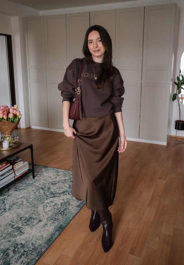 Donna in piedi all'interno che indossa un maglione marrone scuro con scritta, gonna in raso abbinata, stivaletti e una borsa a tracolla bordeaux.