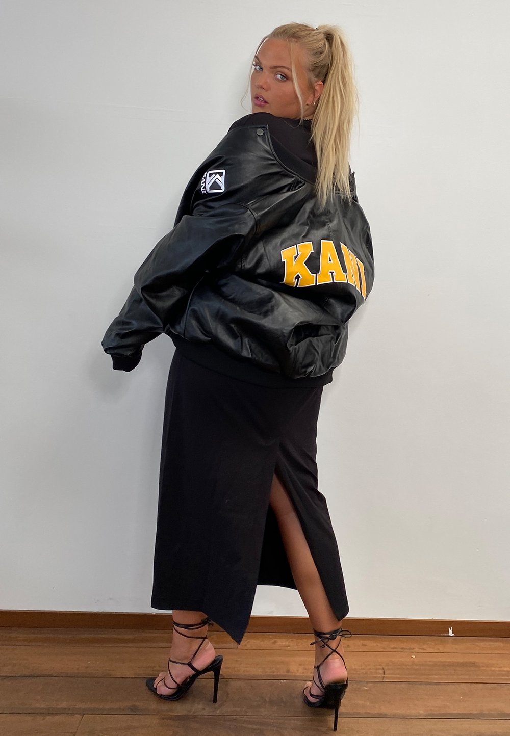 Must musta nahkavest, millel on seljas kollane 'KANI' kiri, kantud sileda musta kleidi peal, millel on reie kõrge lõhik ja mustad rihmaga kontsad.