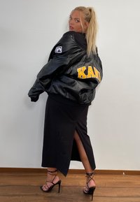 Must musta nahkavest, millel on seljas kollane 'KANI' kiri, kantud sileda musta kleidi peal, millel on reie kõrge lõhik ja mustad rihmaga kontsad.