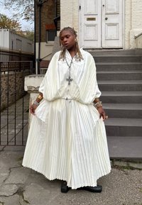 Hvid plisseret kjole med høj krave og brede ærmer. Har snøre i taljen, lagdelt tekstur og et korshalskæde. Sorte sko.